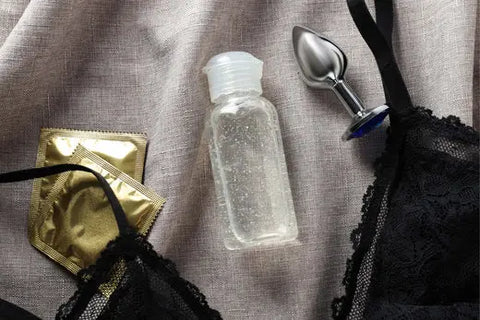 Image of some lube, two condoms, black lacy lingerie, and a butt plug on a bed - LoveKinx UK